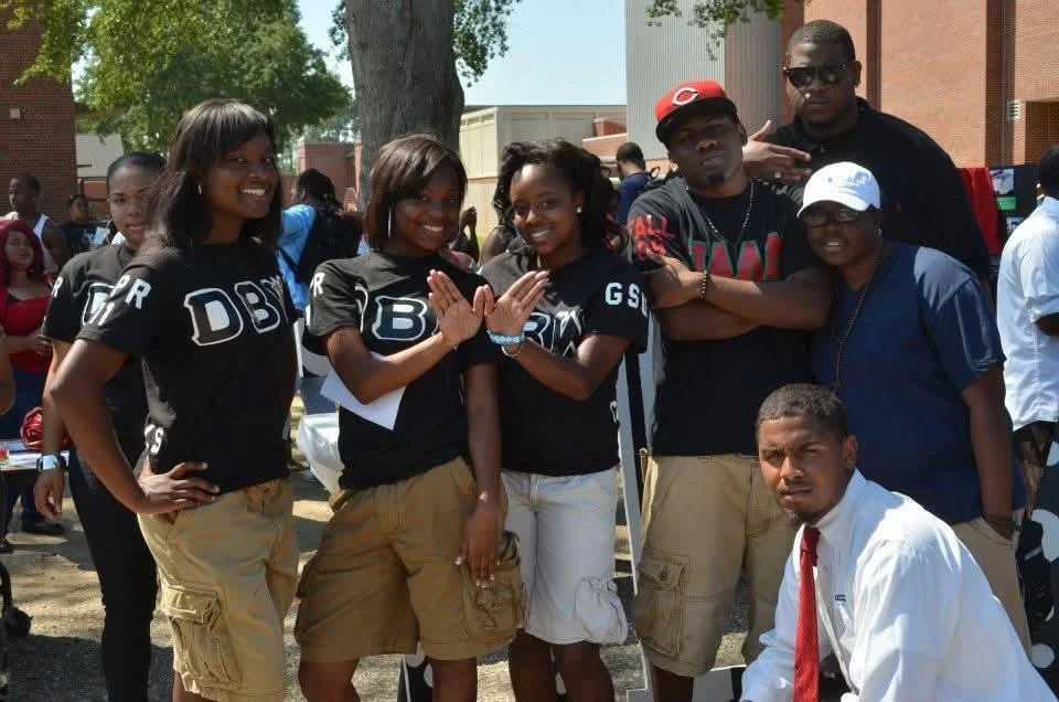 group of young African American people, smiling, posing outdoors, some making peace signs or crossing arms, with a few wearing matching black shirts with "DBR" logos, and one person in a white shirt and red tie.
