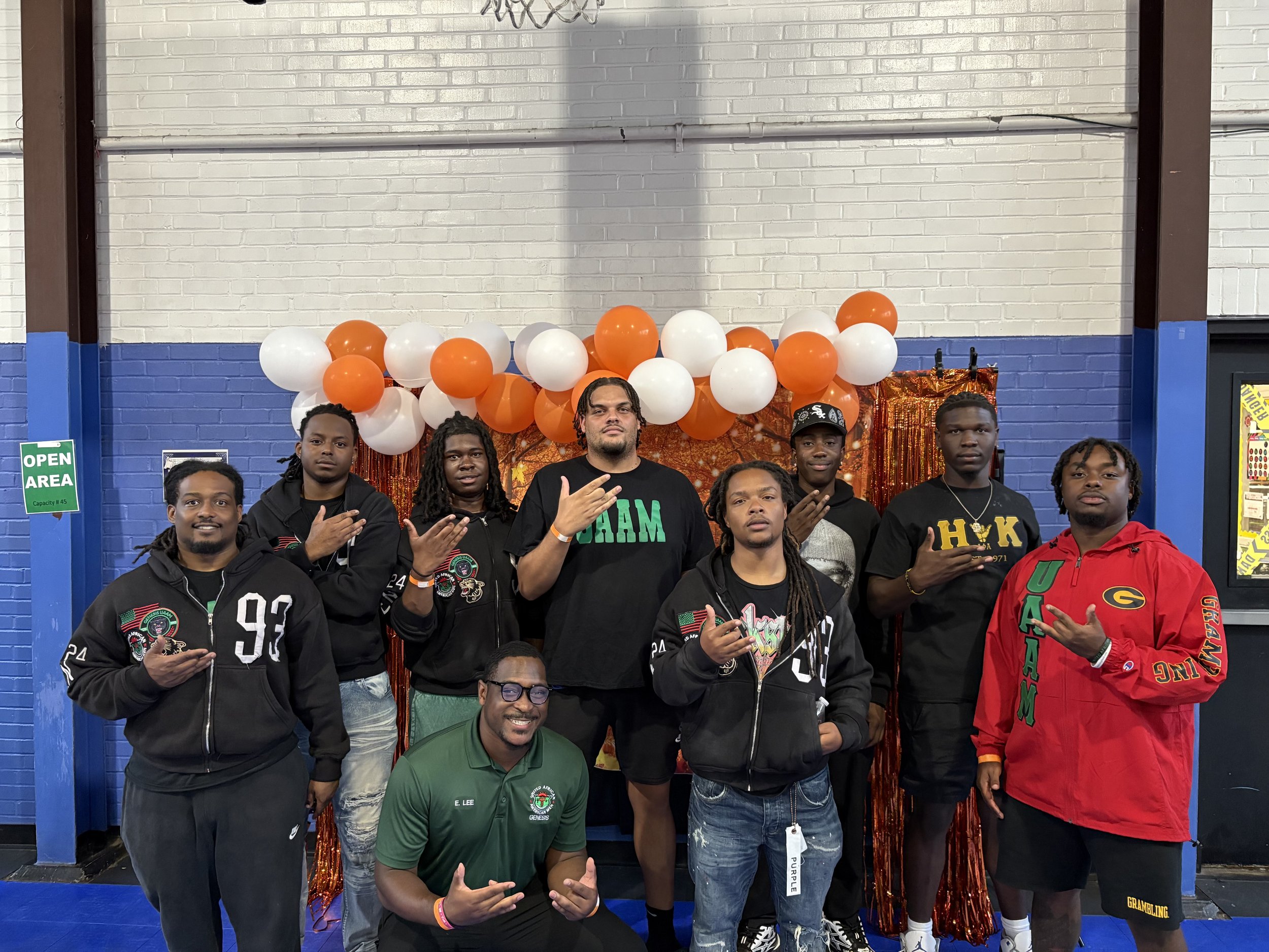 Group of nine young men and one adult male posing in front of a balloon arch with orange and white balloons and a shiny orange backdrop. They are wearing casual outfits, some with branded sportswear or hoodies, and making hand gestures.