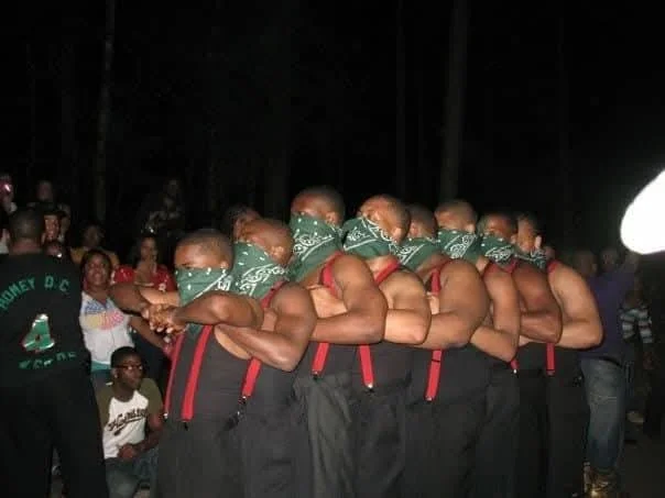 A group of men with green bandanas covering their mouths, standing in a line with their arms crossed over their chests, at night with an audience behind them.