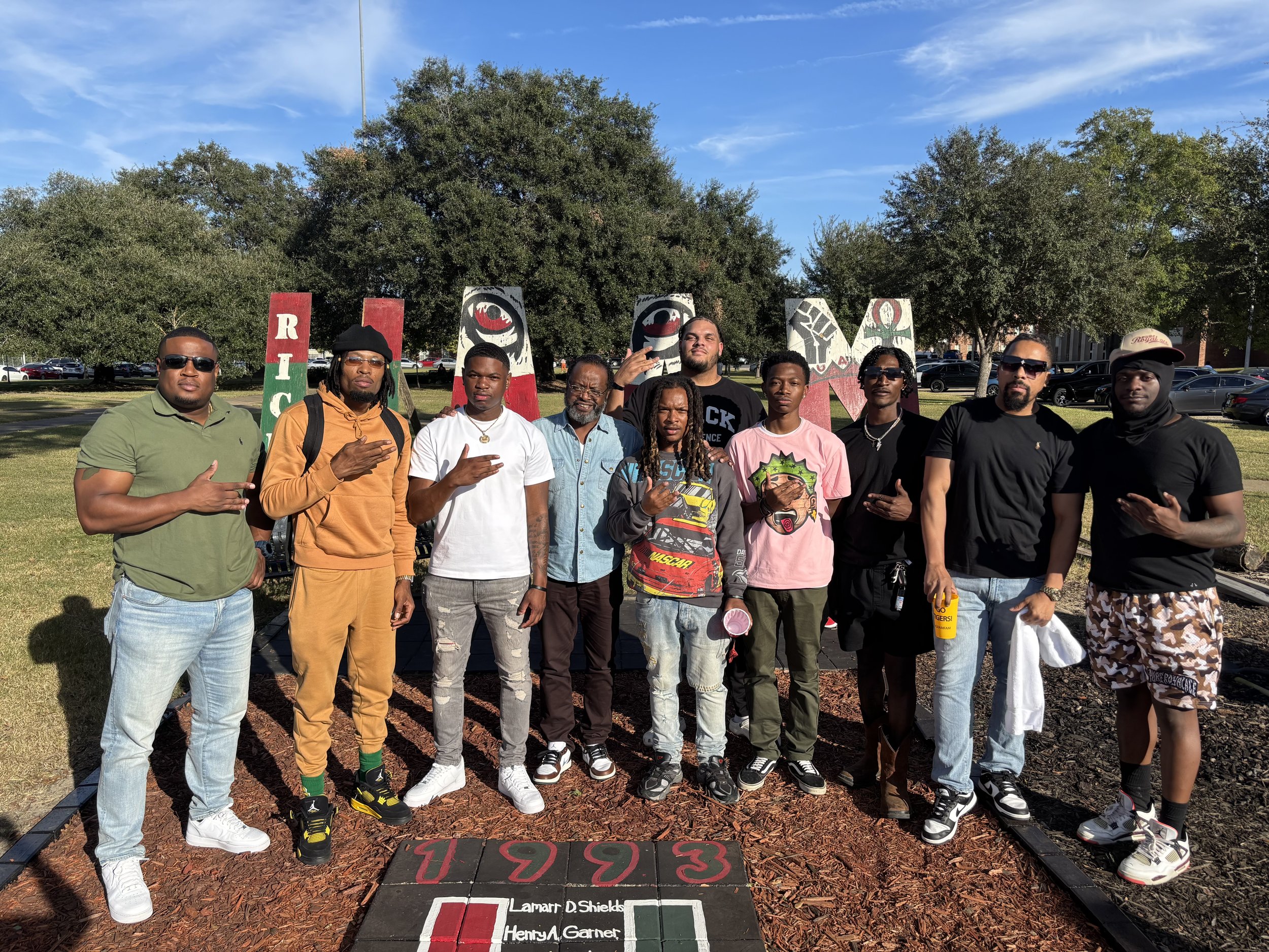 Group of eleven young men standing outdoors on a sunny day, posing in front of a decorative sign with trees and parked cars in the background, some making hand gestures.