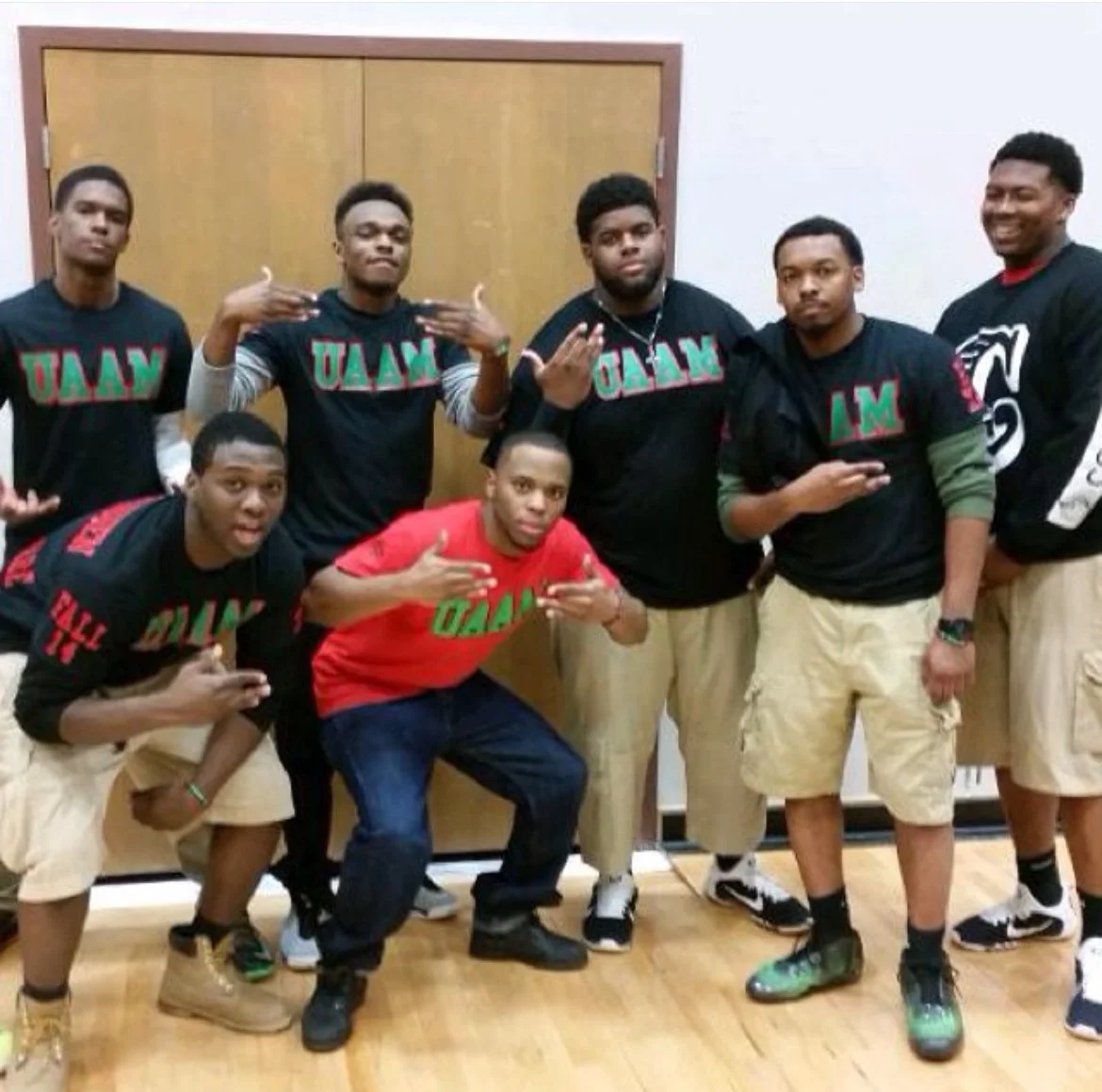 Group of eight young men posing together indoors, wearing matching t-shirts with the text 'UAN,' some making gestures, standing in front of a wooden door with a white wall.