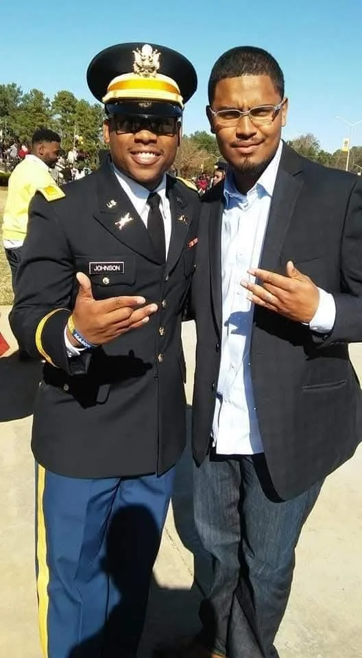 A man in a U.S. military dress uniform with a hat and sunglasses, and another man in a dark suit and light shirt, standing outdoors with trees in the background. They are smiling and making hand gestures, possibly at a graduation or special event.