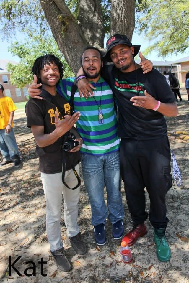 Three men smiling and posing together outdoors under a tree. Two of them are making hand signs, and one has an arm around another. There are other people in the background.
