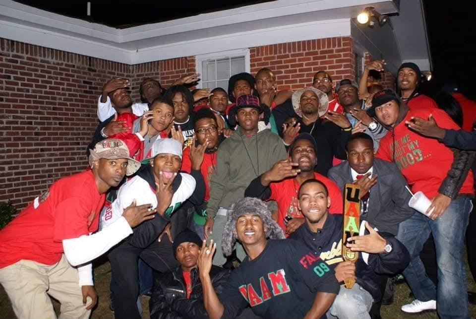 Group of young men posing together outside a brick building at night, some making hand gestures, celebrating a graduation or achievement.