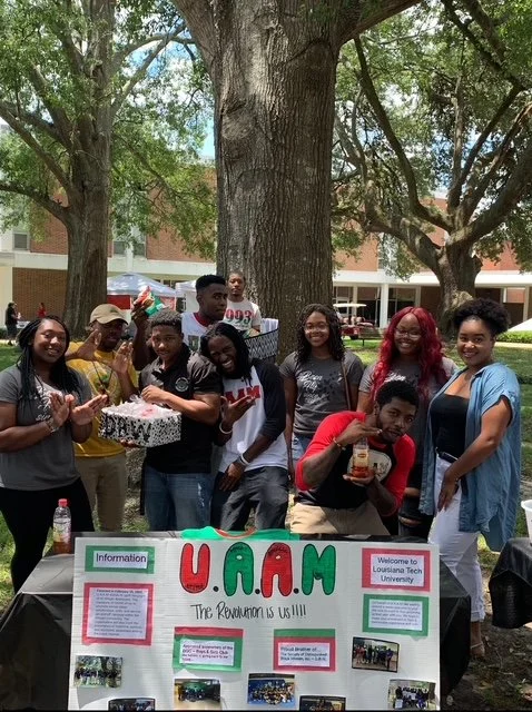 Group of young people in casual clothing gathered outdoors around a table with an informational poster for U.A.A.M, standing in front of large trees and a building.