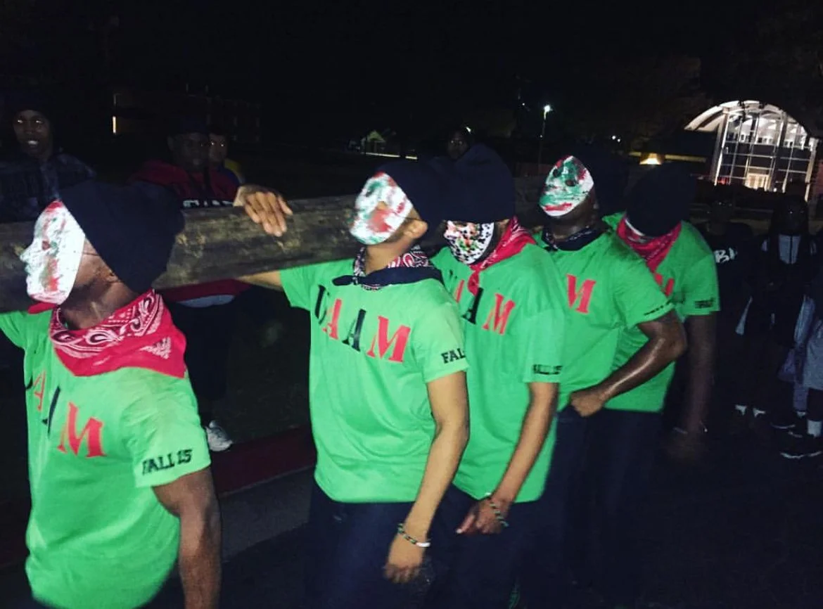 Young men wearing green shirts, bandanas, and masks carrying a wooden cross during night event.