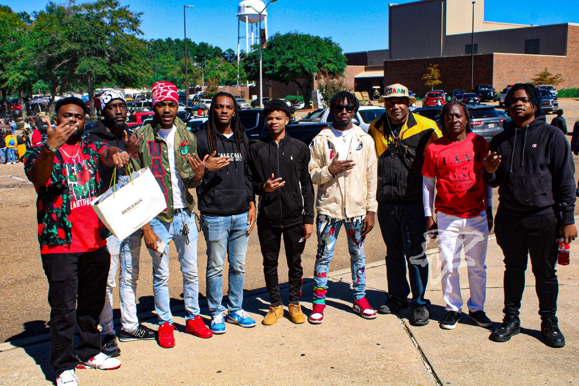 Group of ten young men standing outside in a parking lot, posing for a photo, with a large building, trees, and parked cars in the background.