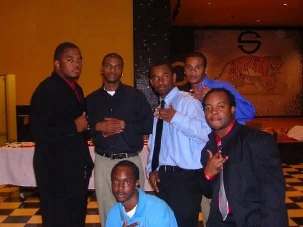 Six men posing together at an indoor event, some making gestures or holding objects, with a tiger mural in the background.