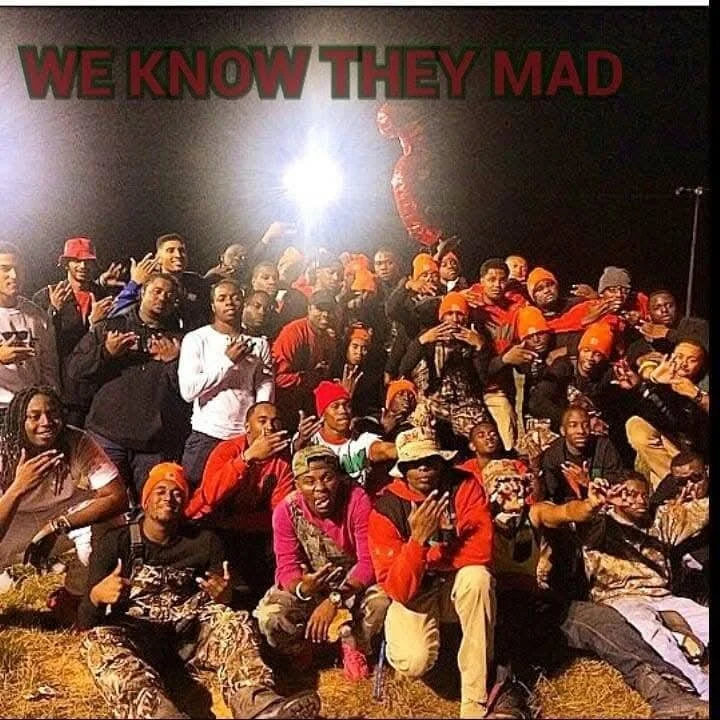 A group of people gathered outdoors at night, some making hand gestures or holding their hands over their mouths, with a large red and green sign above them that reads 'WE KNOW THEY MAD'.