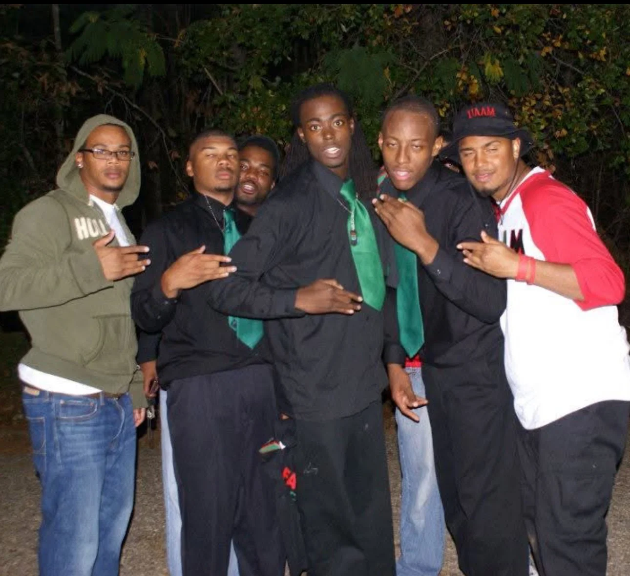 Group of six young men standing outdoors at night, some making hand signs, with a background of dark trees.