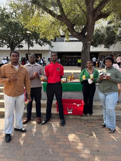 Group of five young adults standing outdoors near a tree, holding snacks and drinks, with a food stand behind them and other people in the background.