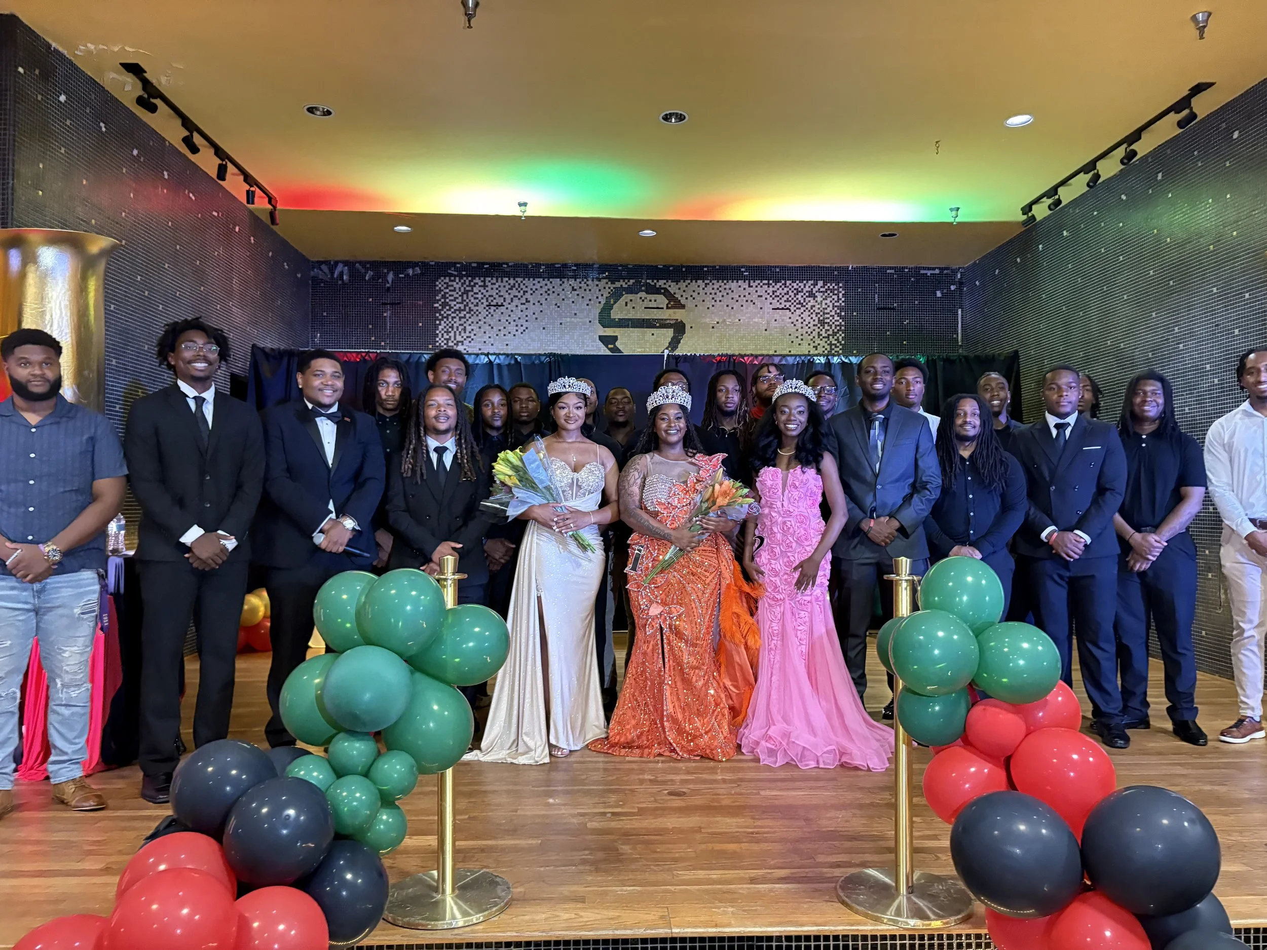 A group of people dressed in formal attire standing on a stage decorated with colorful balloons, with two women in the center wearing crowns and holding bouquets of flowers. The background features a black curtain and a decorative wall with a logo.