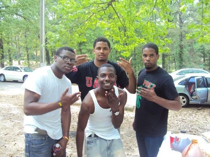 Four young men outdoors in a wooded area, posing for a photo. They are casually dressed, with one in a white tank top and others in black t-shirts. One man is smiling and making a gesture with his hand, others making hand signs. Cars are visible in t
