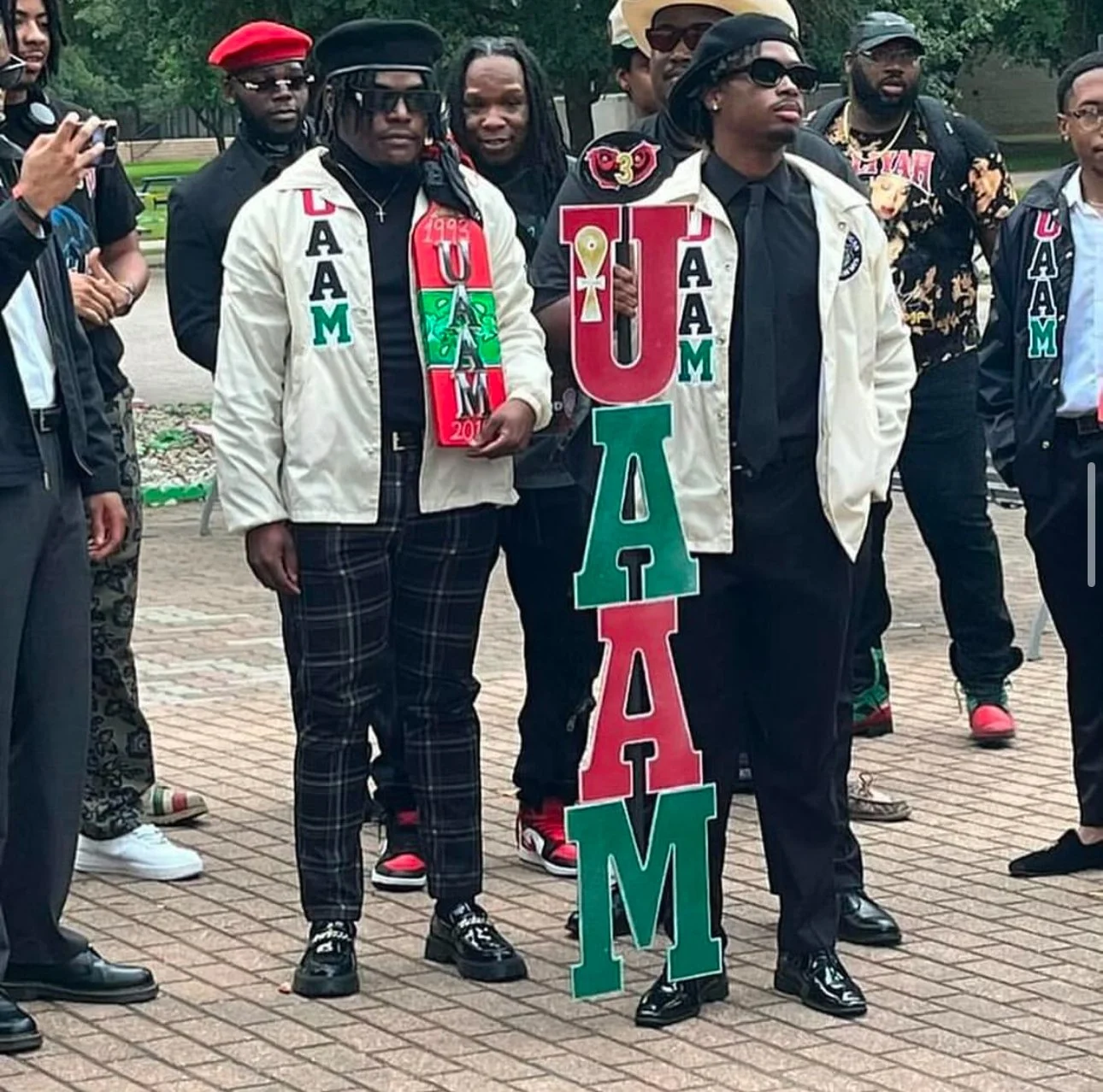 Group of people at a gathering, holding a large colorful sign spelling 'UAM' in red, green, and black letters, with some individuals wearing casual and fashionable attire and sunglasses.