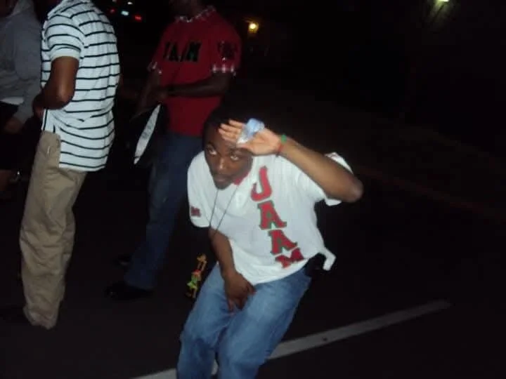 A young man in a white T-shirt with red letters spelling 'JAMA' standing on a street at night, saluting the camera with his right hand, while posing in a playful manner.