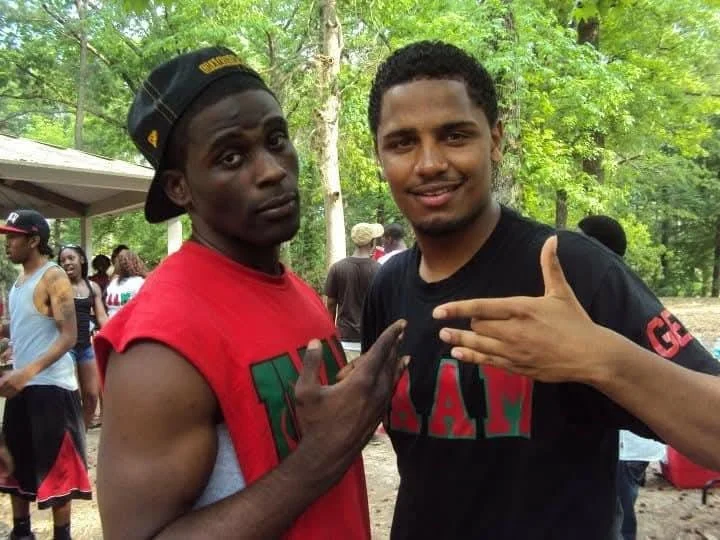 Two men standing outdoors in a park with green trees in the background. One man wears a red sleeveless shirt and a black cap worn backwards, while the other wears a black t-shirt. They are making hand gestures and smiling at the camera.