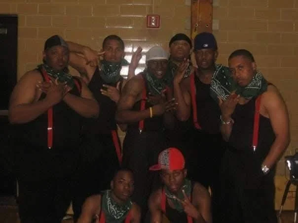 A group of eight men posing indoors, some making hand gestures, wearing bandanas and sleeveless shirts, with a brick wall in the background.