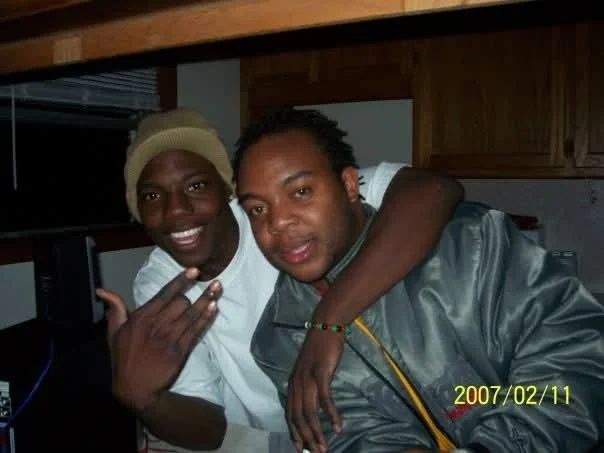 Two men smiling and sitting closely together, one with his arm around the other, in a room with wooden cabinets. The man on the left is wearing a beige cap and making a hand gesture, the man on the right is wearing a gray jacket. The date "2007/02/11