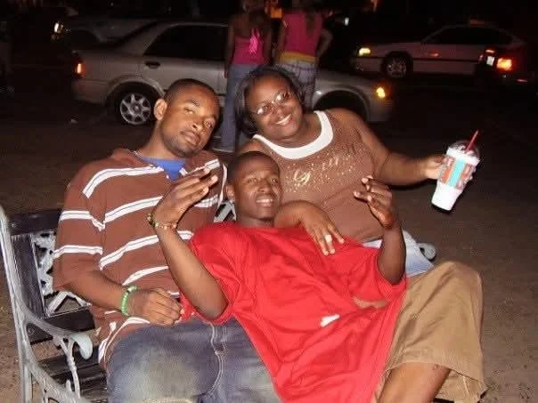 Three people sitting on a park bench at night, smiling and posing for the camera. Two men and one woman, with cars in the background.