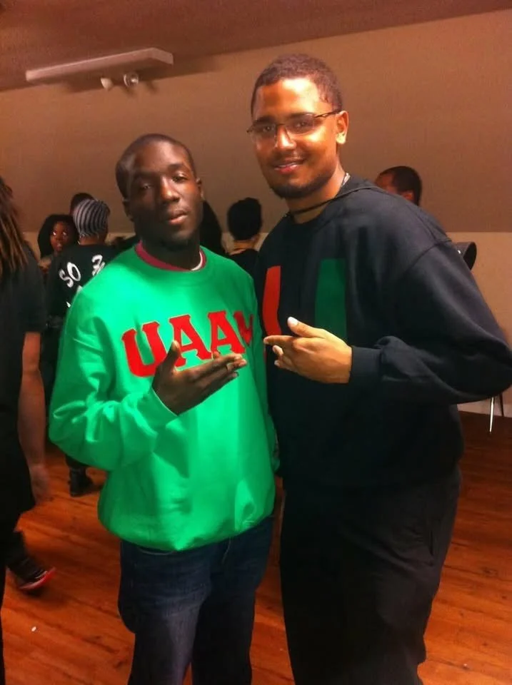 Two men standing close together at an indoor event, both making hand gestures and smiling at the camera. The man on the left is wearing a bright green sweatshirt with red Greek letters and the man on the right is wearing a black hoodie with a colored