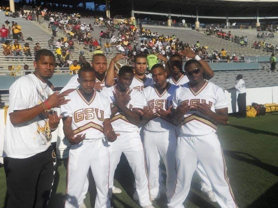 Group of young men in white football uniforms with 'GSU' on the chest, posing on a football field with bleachers and spectators in the background.