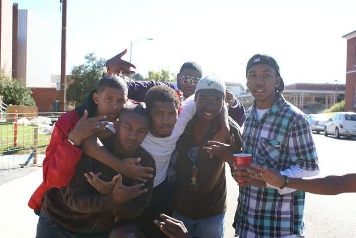 Group of six young men outdoors on a sunny day, some making hand gestures, one holding a red cup, standing on a street with parked cars and buildings in the background.