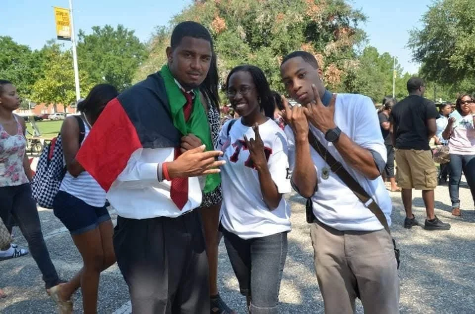 Three young people standing outdoors, posing and making hand gestures. The person on the left has a flag draped over their shoulders. There are other people and trees in the background.