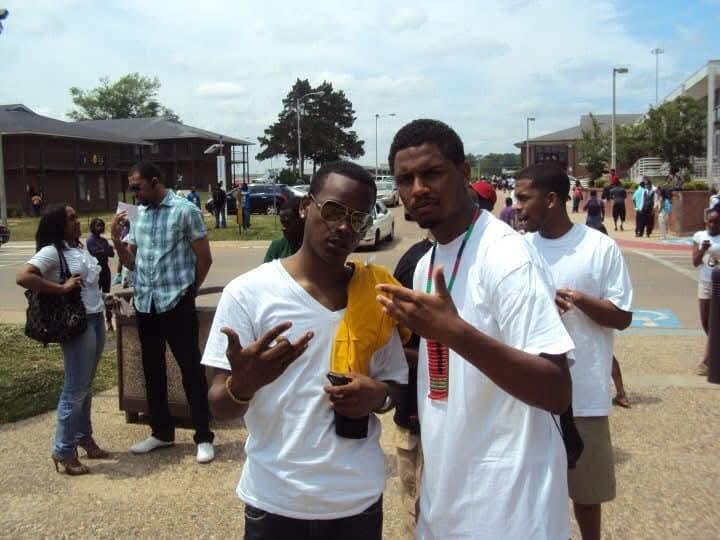 Two young men posing for a photo outdoors at an event. One is wearing sunglasses and a white T-shirt, holding a phone, making a hand gesture. The other is also in a white T-shirt, looking at a phone, with a medal around his neck. People are gathered 