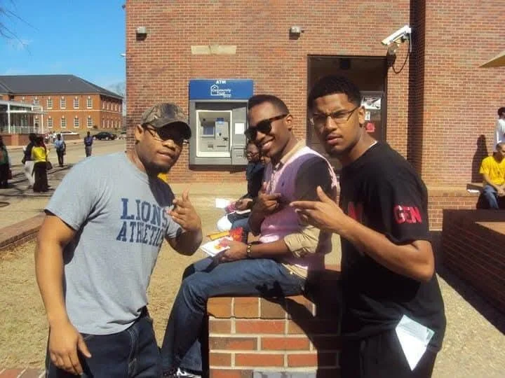 Three young men posing for a photo outside a brick building, with one sitting on a brick ledge and the other two standing on either side, all making hand gestures.