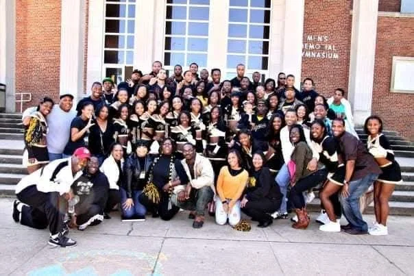 Group of students and teachers gathered outside a school building with large windows and steps, posing for a class photo.