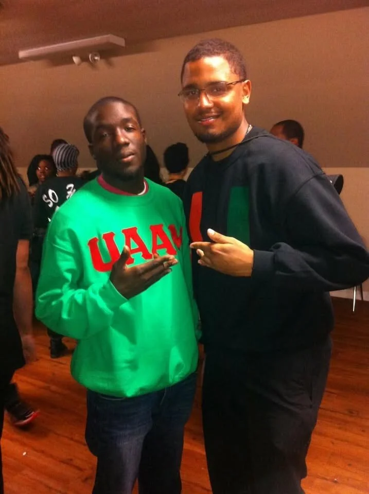 Two men posing together indoors, one wearing a green sweatshirt with red letters, the other wearing a black sweatshirt with a colorful design, both making hand signs and smiling.