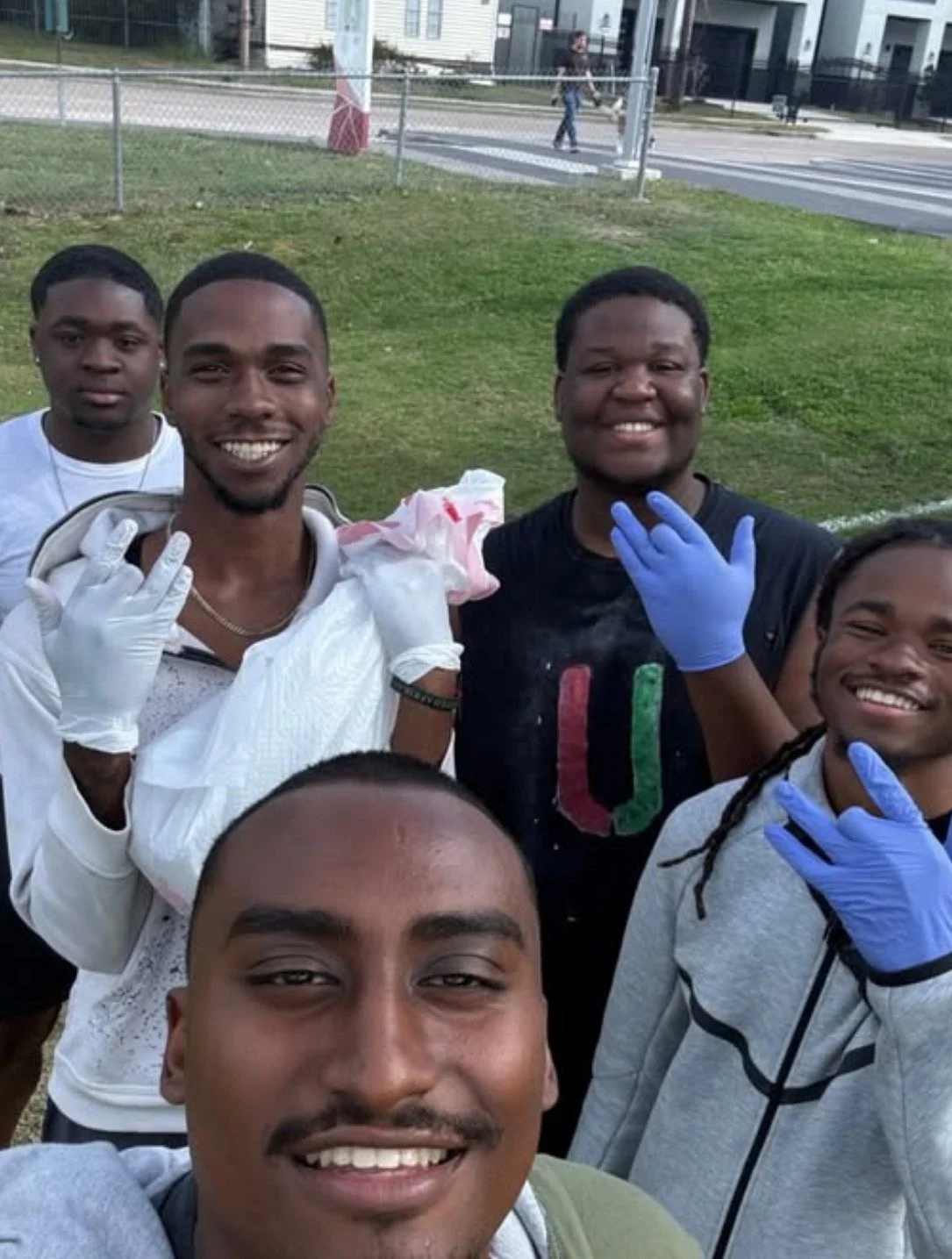 Group of five young men smiling and posing for a photo in an outdoor park area, some wearing gloves, with grassy field and street in background.