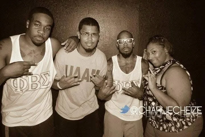 Four people standing together, three men and one woman, posing for the photo. The men are wearing tank tops with Greek letters, and the woman has her hand on one man's shoulder. The background is plain and dark.