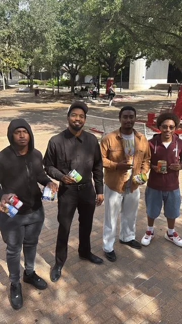 Four young men standing outdoors in a park, holding bottled drinks and snacks, with trees and other people in the background.