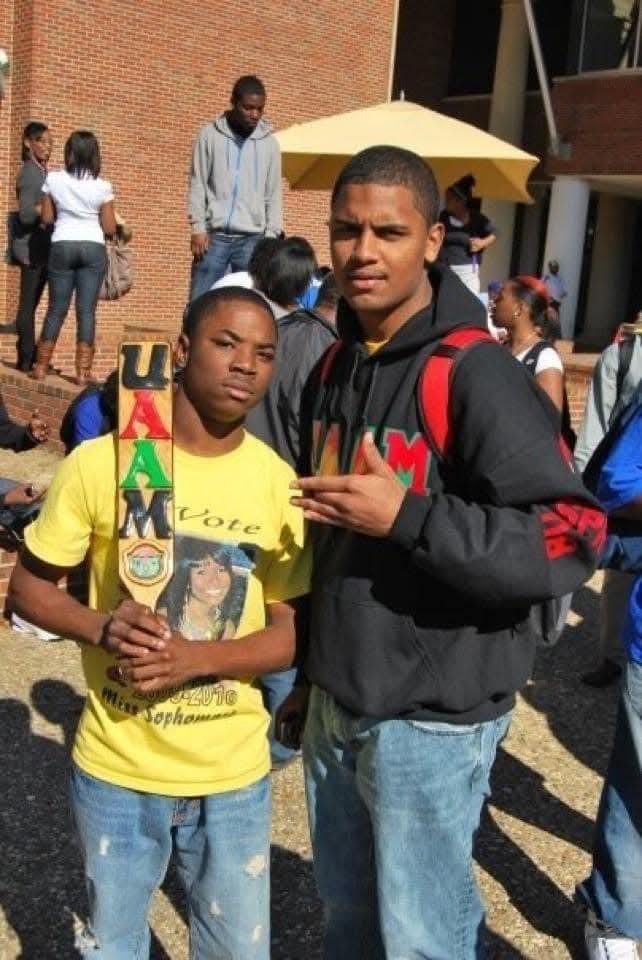 Two teenagers, a girl and a boy, standing together outdoors during the day among a crowd of students. The girl is holding a large medal and wearing a yellow T-shirt with a portrait and text, while the boy is dressed in a black hoodie and blue jeans. 