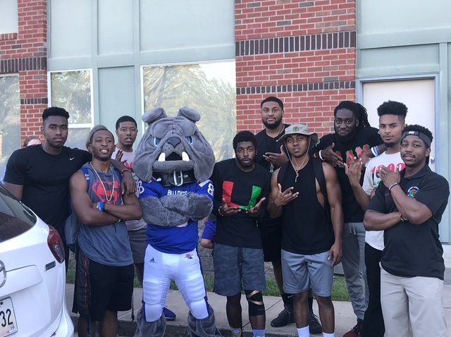 A group of nine young men and a bulldog mascot posing outdoors in front of a brick building. The mascot is in a sports jersey, and the men are dressed casually, some making hand gestures.
