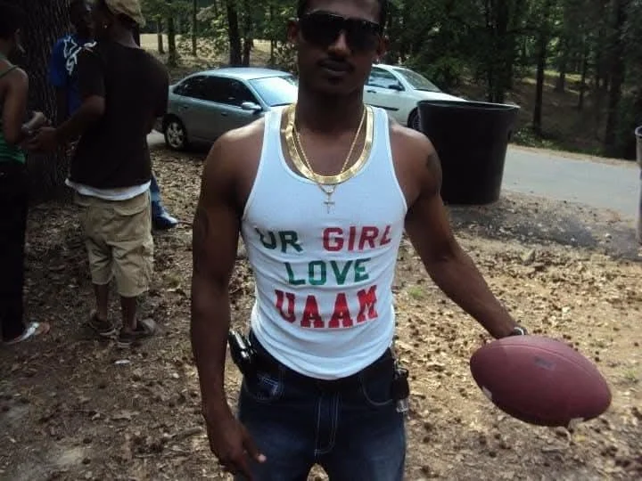 A young man holding a football outdoors, wearing sunglasses, a white tank top with colorful text, gold necklaces, and jeans. There are other people and cars in the background.