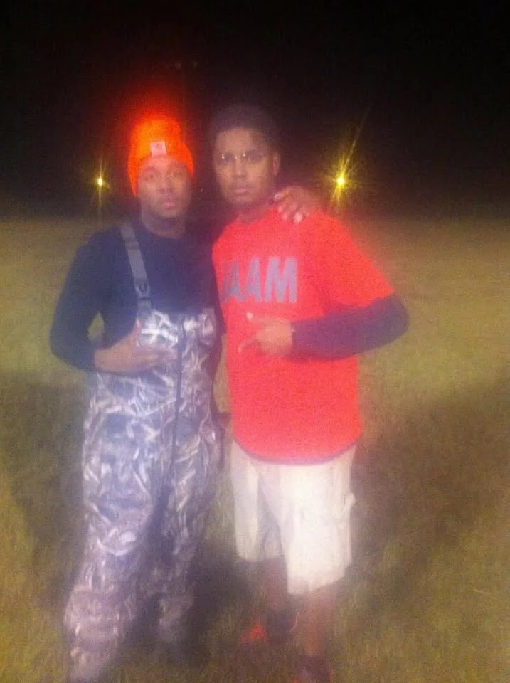 Two men standing outdoors at night, one wearing a camouflage outfit and a bright orange hat with a headlamp, the other in a red Miami shirt and beige shorts, with their arms around each other.