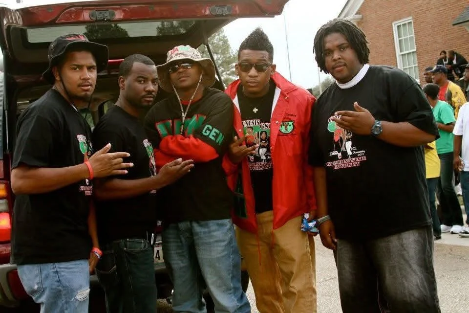 Five young men standing outdoors near a vehicle, posing for the photo. They are wearing casual clothing, some with graphics or logos on their shirts.