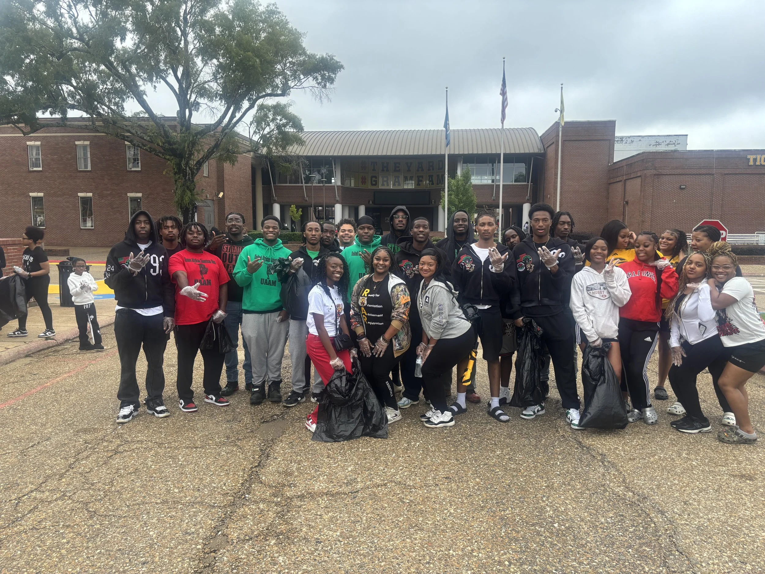 A group of young people posing outside a school building, some holding trash bags and wearing gloves during a community cleanup event.