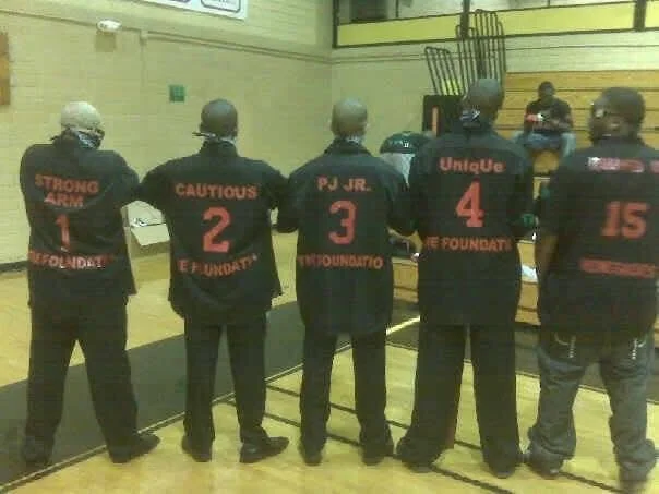 Five individuals with painted numbers and words on their backs, standing in a gymnasium, facing away from the camera.