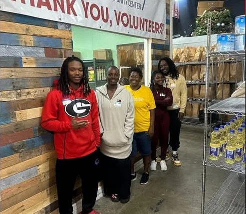 Five people standing in a row inside a community center or food pantry, with shelves of food and bottled water nearby. A sign above them reads 'Thank you, volunteers'.