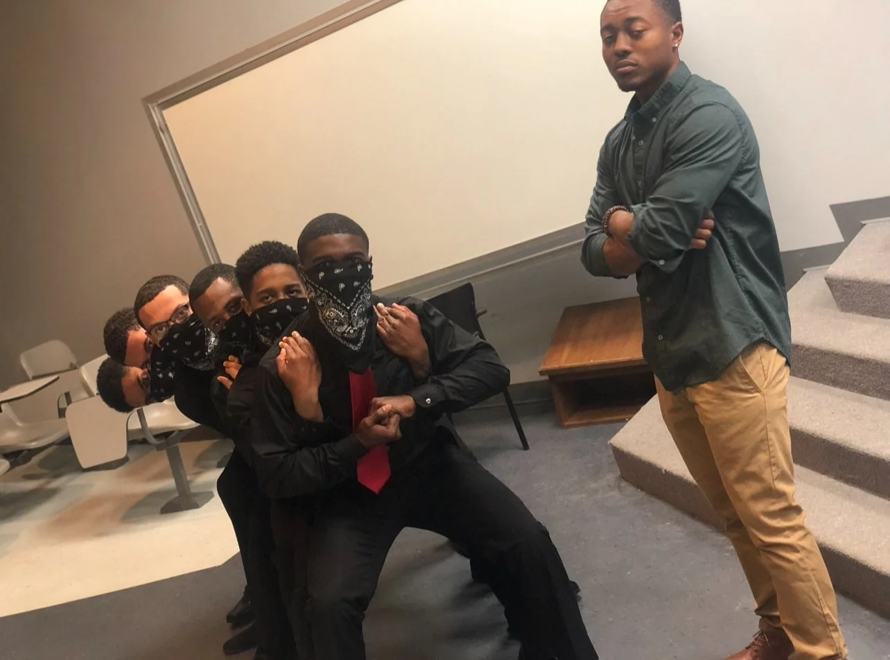 A group of five men wearing black shirts, black bandanas over their faces, and sunglasses, are in a fighting stance in a room. They are pretending to be tough and serious. Standing opposite them is a man in a green shirt and tan pants, standing with 