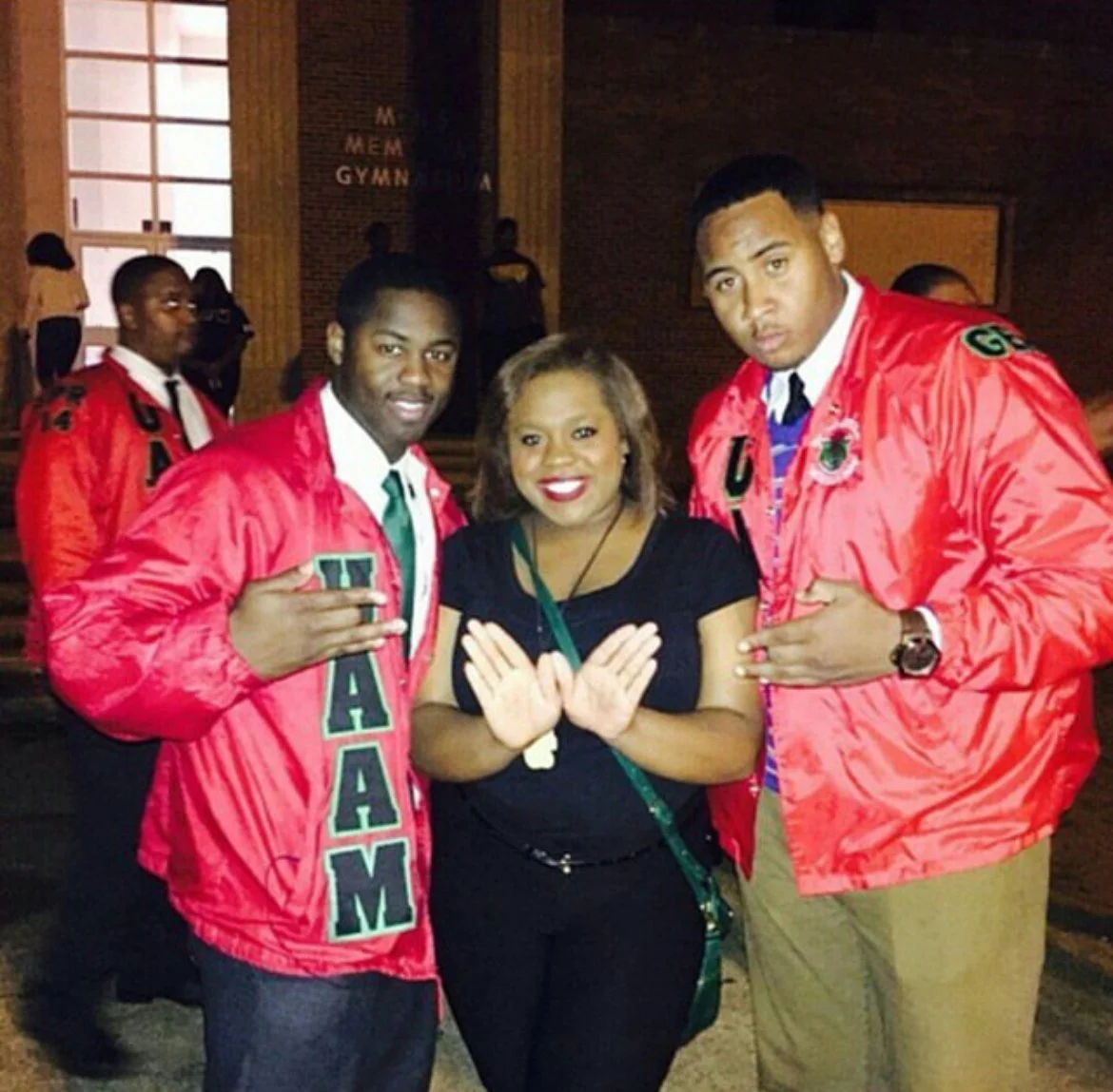 Two young men wearing red jackets with green letters and a woman in a black top in between, with the woman making a gesture with her hands. The background shows others in a gymnasium or auditorium setting.