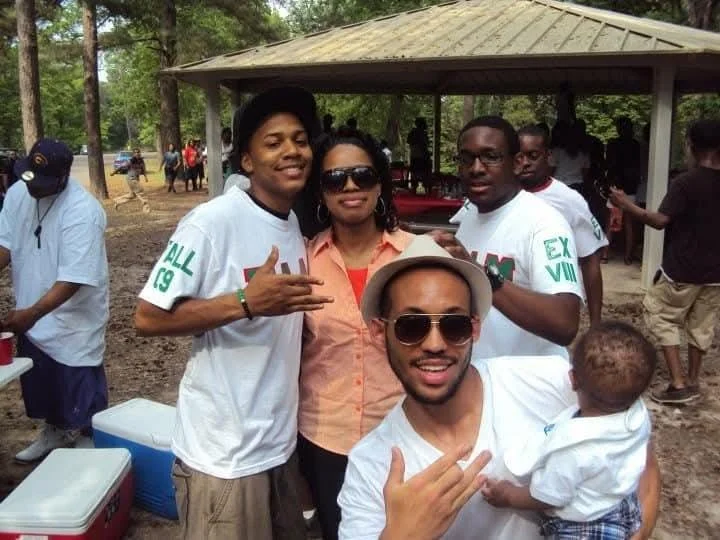 Group of five people, including a woman and a young man, posing outdoors at a picnic area with trees and a pavilion in the background.