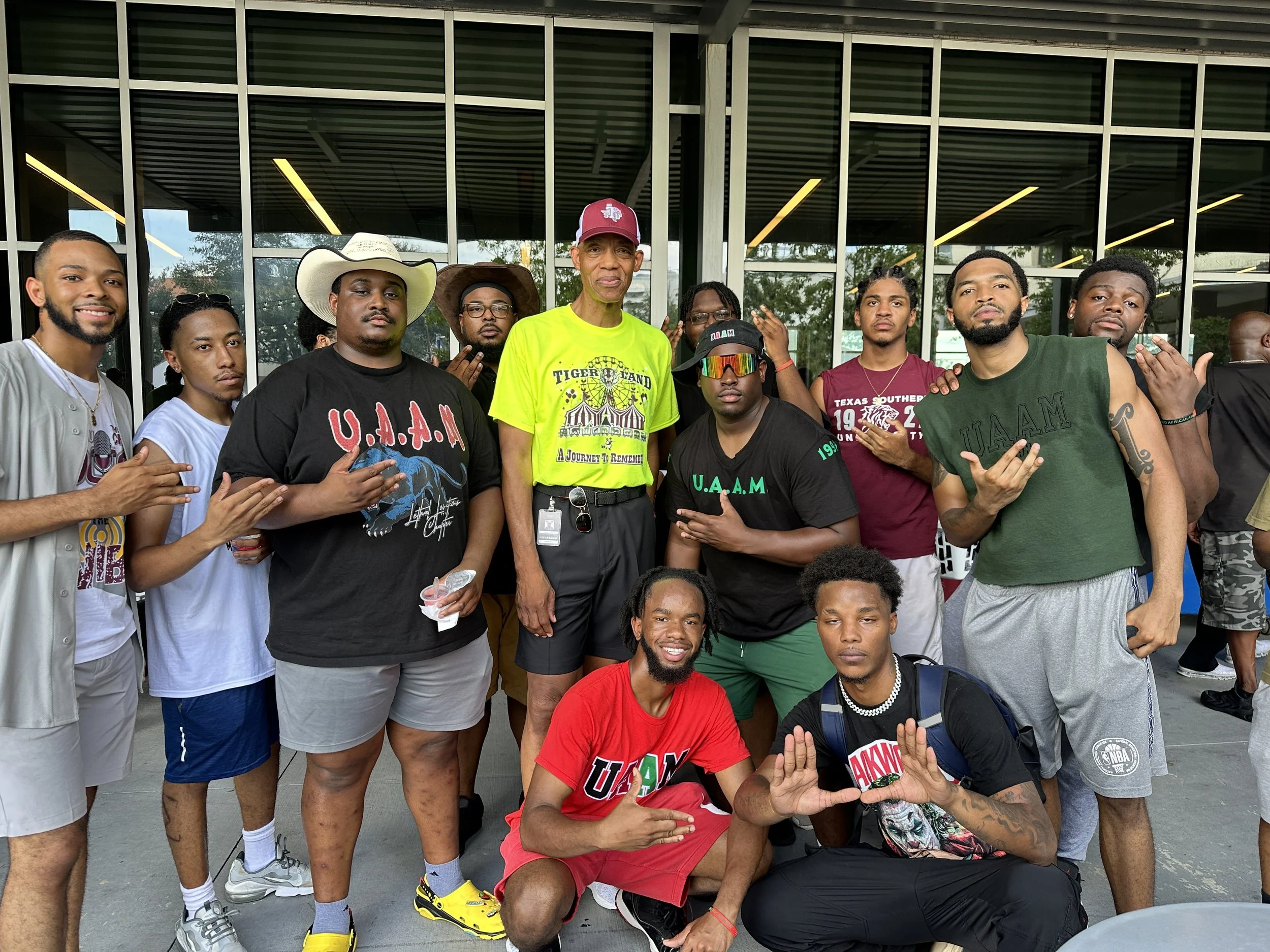 Group of young men and one older man posing outdoors in front of a building with glass windows, some wearing casual clothes, hats, and sunglasses, making hand gestures and smiling.