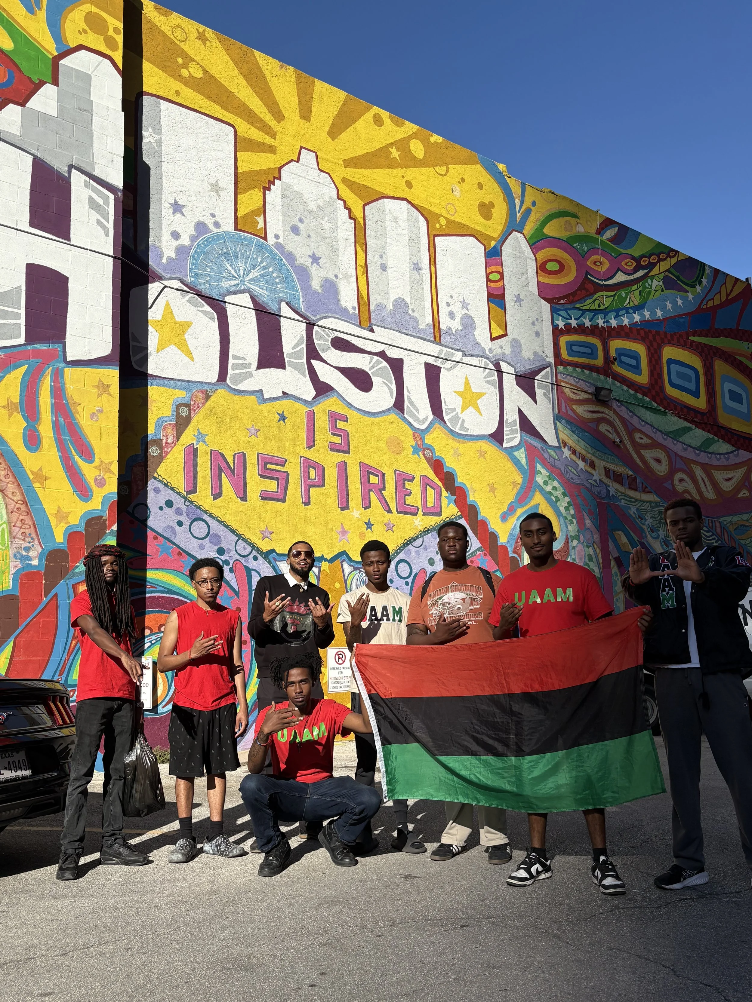 A group of seven young men posing for a photo holding a flag with red, black, and green stripes in front of a colorful mural with the text "Houston is Inspired." The mural features bright yellow, purple, and blue colors, cityscape outlines, stars, and abstract patterns.