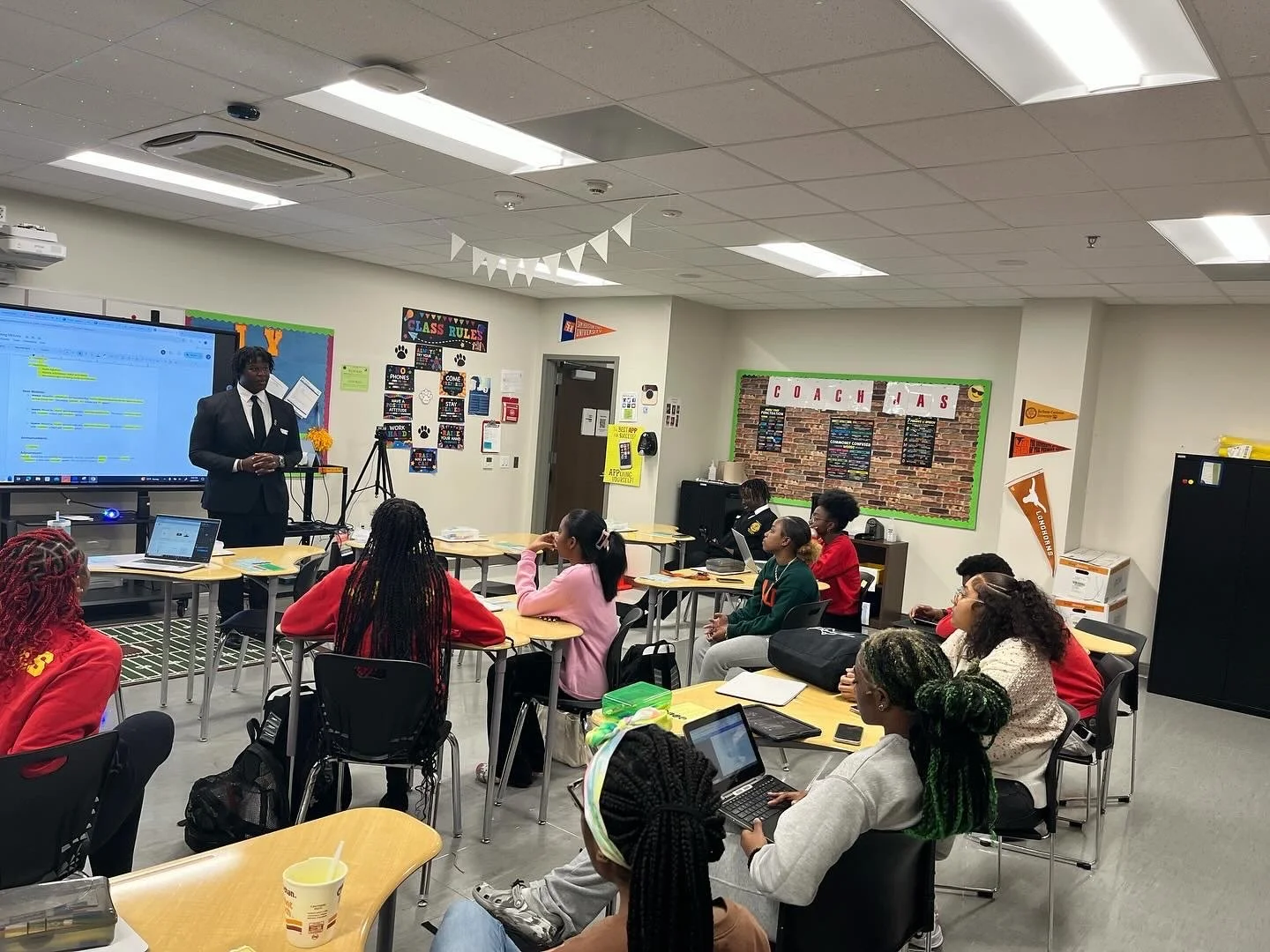 A classroom with a person at the front giving a presentation to students seated at tables, some taking notes or using laptops. The classroom is decorated with posters, banners, and conference banners.