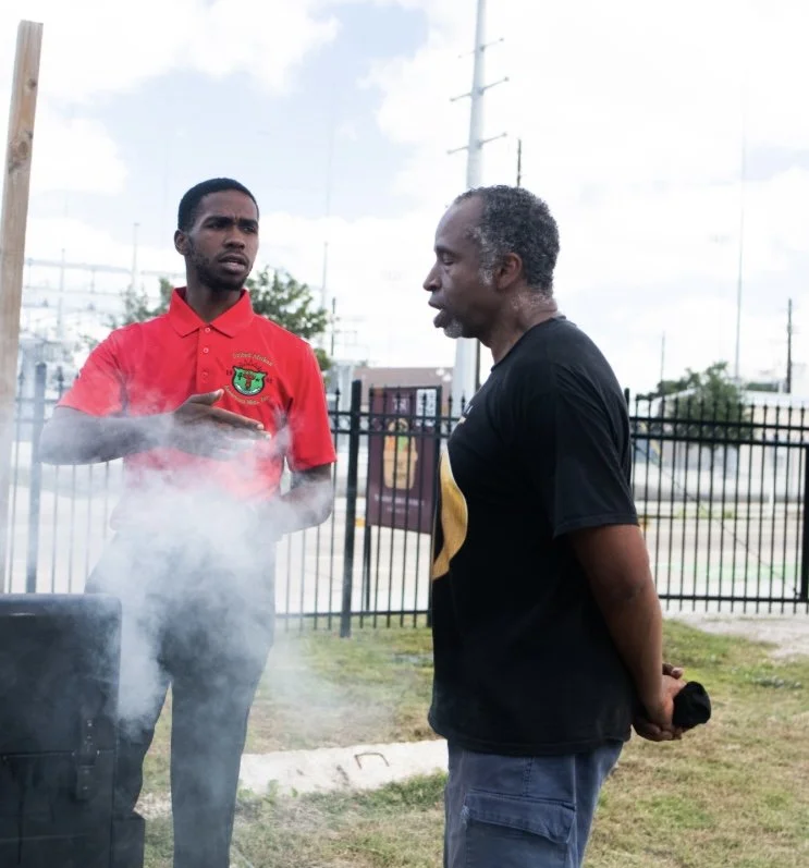 Two men having a conversation outdoors near a fence, with one man gesturing with his hand, smoke rising from a grill or smoker nearby.
