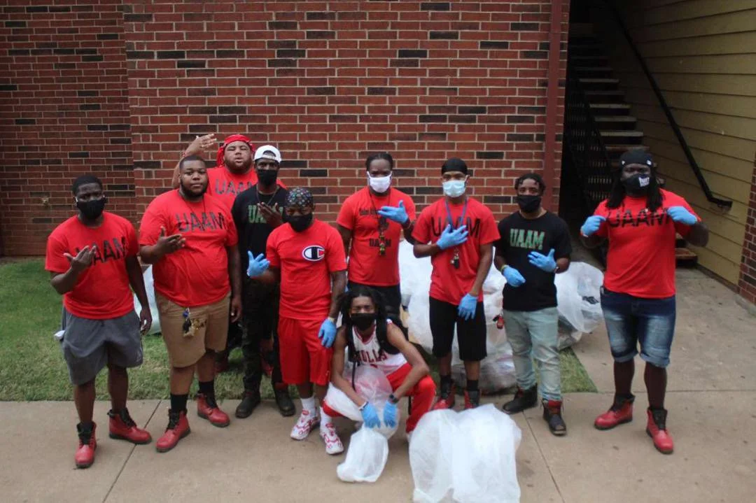 A group of nine young people, dressed in red and black shirts with 'UM' or 'UMM' printed on them, pose outdoors in front of a brick wall and a stairway. They are wearing face masks and gloves, and some are making hand gestures. There are large trash bags filled with collected items on the ground.